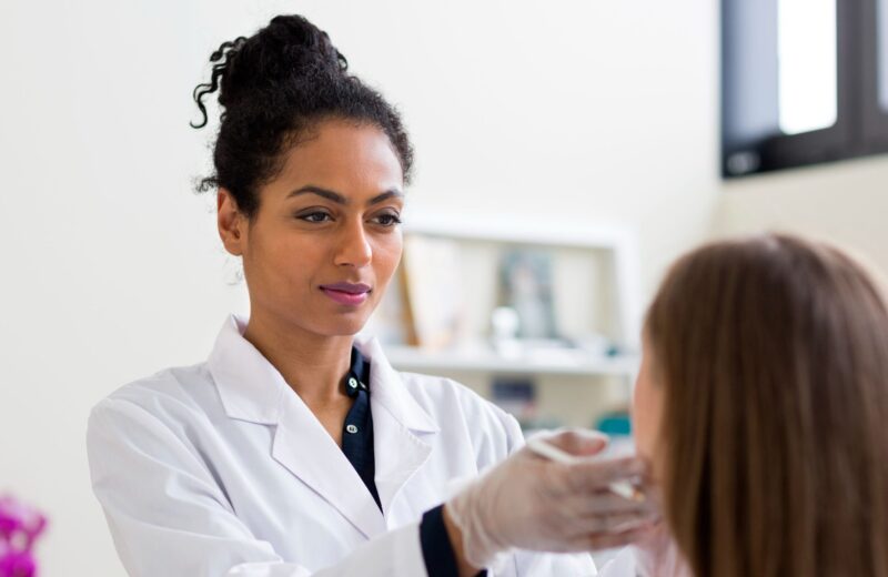 A clinician wearing a white coat and gloves examines a patient’s face during a consultation.